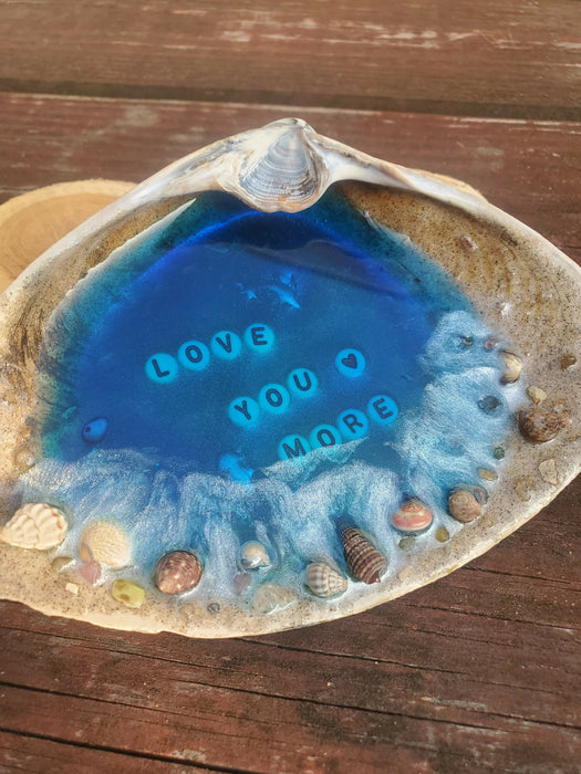 A natural clamshell repurposed into an ocean blue resin Beach Scene set right inside the center of the shell.  It has tiny shells, Sand, and small Gemstones and has Love you more written with white and black letter beads in the center of the blue resin Ocean water.