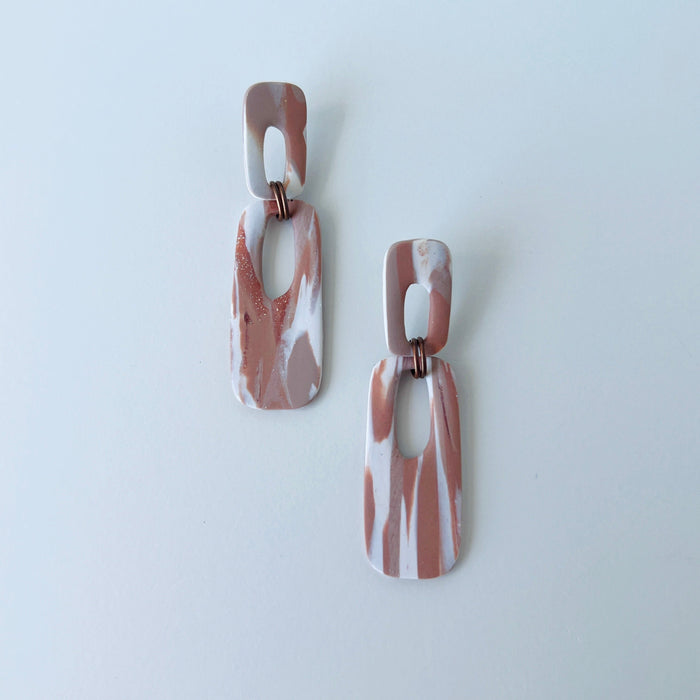 A pair of handmade rectangular drop earrings with a cutout center, made from marbled polymer clay in shades of terracotta brown, shimmering copper, and white. The top and bottom segments are joined with antique copper-colored jump rings. The earrings have embedded titanium posts.