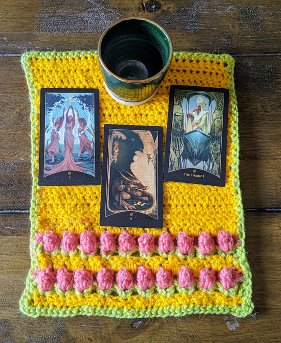 Photograph of a small, yellow, crocheted altar cloth with two rows of pink tulip stitches at the bottom, with a lime green border. There are three tarot cards and a small ceramic cup on it.