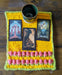 Photograph of a small, yellow, crocheted altar cloth with two rows of pink tulip stitches at the bottom, with a lime green border. There are three tarot cards and a small ceramic cup on it.