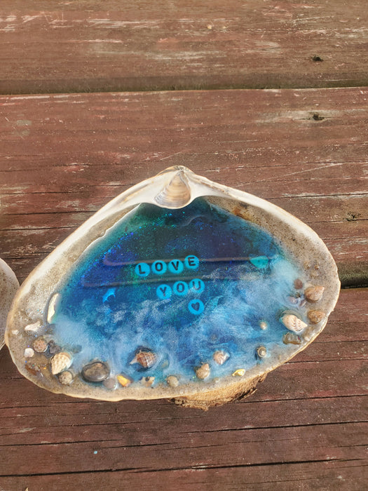 This is another view of the large clamshells that acts as the base of the bright blue ocean resin Beach Scene set inside the center of the shell.  There are white waves at the sandy shore line area as well as tiny shells and Gemstones.  This shell says Love You in the center of the blue resin with white and black letter beads and is 1 shell in this set of 2.