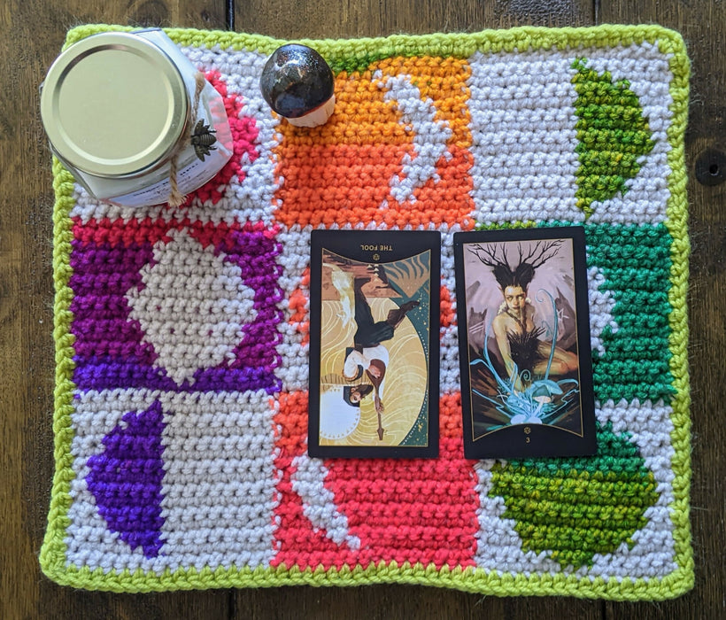 Photograph of a tarot cloth with a checkerboard moonphases design made with rainbow and white yarns with a lime green border. There are two tarot cards, a candle, and a ceramic mushroom on it.
