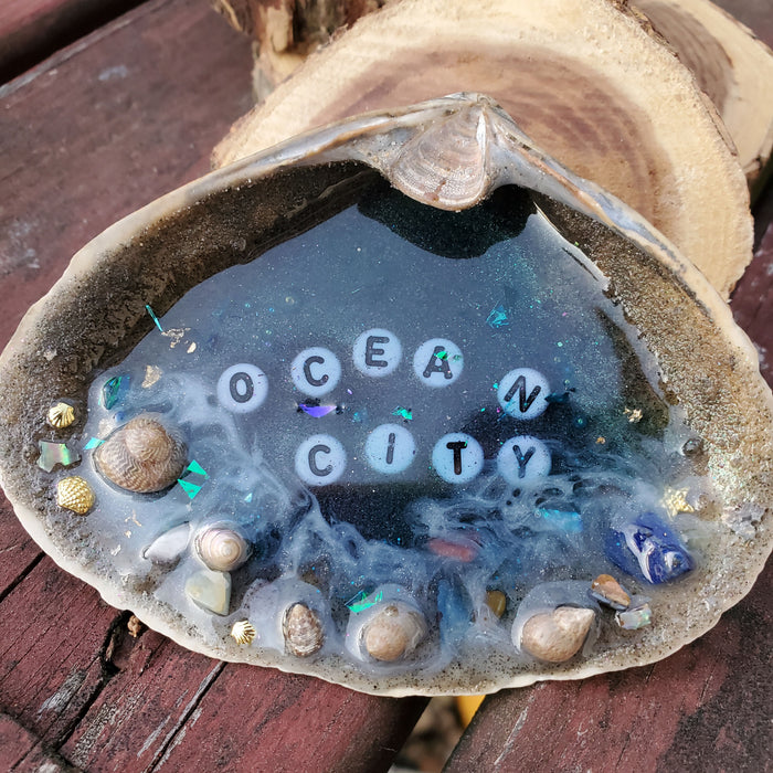 A white clam Shell with blue resin in the center making a resin Ocean scene.  There is sand on the outer edge of the shell with Ocean City in the center in white and black letters beads