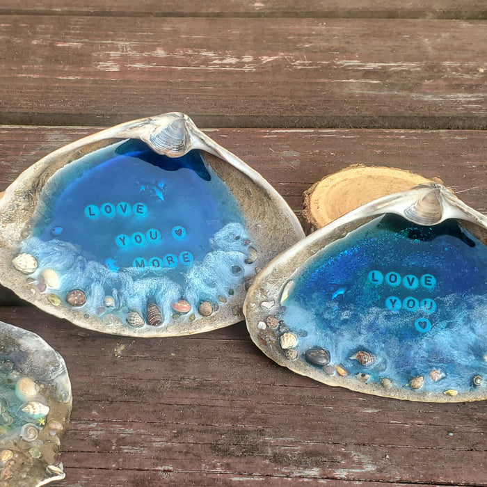 This shows the set of 2 Love You and Love You More resin ocean Beach scenes set in the center of large natural Clam shells.  The resin in the center is bright blue to act as the water and there is Sand along the outer edge to represent the shore line.