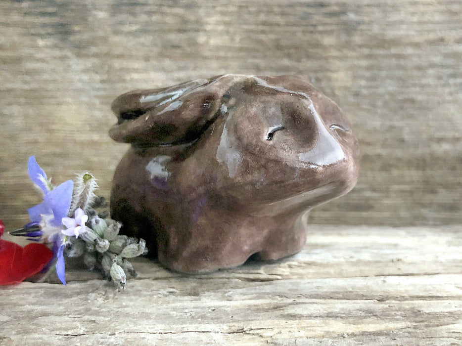 Raku ceramic sculpture of a rabbit. It is sitting with its long ears folded down its back and is glazed in dark, warm brown. It has a very sweet, gentle face.