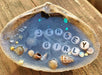 A white clamshell with bright blue resin in the center to make a resin Ocean Beach scene inside the center of the shell.  It has a sandy shore line area with tiny shells on the outer edge of the shell.  Jersey Girl is in the center of the resin "water" with white and black letter beads.