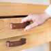 Woman's hand reaching towards a dark brown leather handle on a knotty pine buffet table drawer, which a Harbor Freight woodworking bench.