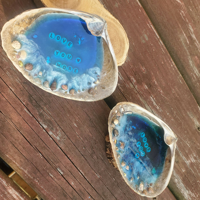 This shows the set of 2 bright ocean blue resin filled beach scenes set inside of natural large clamshells.  There is white at the sandy shore line area on the outer edge of the shells with letter beads inside that says Love You on one and Love Your More on the other one.