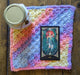 Photograph of a small, square-shaped altar cloth made with blue, purple, orange, yellow, and pink multicolored yarn. There is a candle and a tarot card on it.