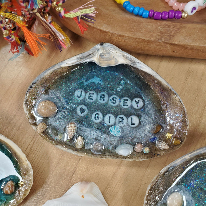 A seashell with a bright blue ocean resin Beach scene in the center and Jersey Girl set inside the blue resin in white and black letter beads.  There is Sand and tiny shells along the outer edge of the shell to act as the shore line.