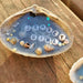 A white clamshell with blue resin ocean scene in the center of the shell, and Jersey Girl in the center in white and black letter beads.  Beads are along the outer edge on the sandy shore line.