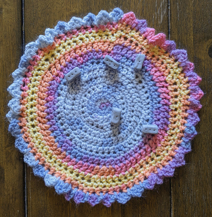 Photograph of a small, round, crocheted altar cloth with ruffled edging. It is made with pastel blue, pink, yellow, and orange multicolored yarn. There is a set of blue knucklebone dice strewn across it.