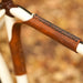 Close-up of a bicycle frame near the seat tube with a brown leather sew-on strap that bridges the top tube and the seat tube against a blurred natural background of fallen leaves
