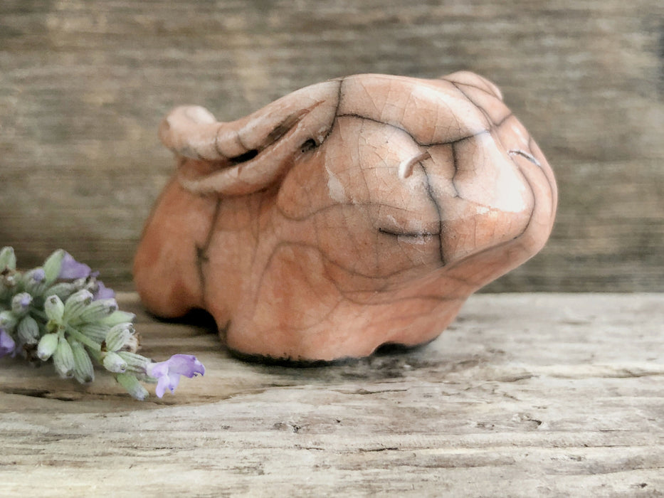 Raku ceramic sculpture of a rabbit. It is sitting with its long ears folded down its back and is glazed in soft, warm peachy orange. It has a very sweet, gentle face.