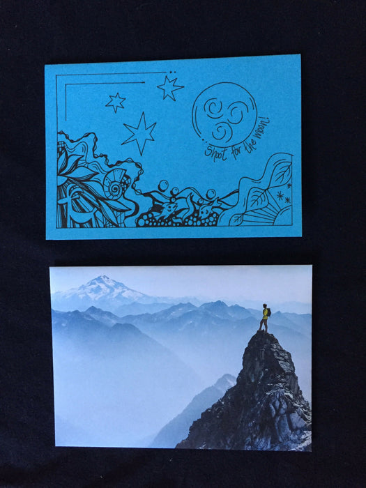 Card features an abstract nature scene including a goat, fish, and a night sky along with the words shoot for the moon. Envelope features photograph of a person standing atop a mountain.
