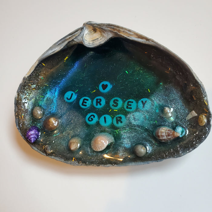 Jersey Girl in white and black letter beads is set inside the center of a natural Clam Shell with blue resin to represent ocean water with sand and tiny shells along the outer edge to act as the shore line.