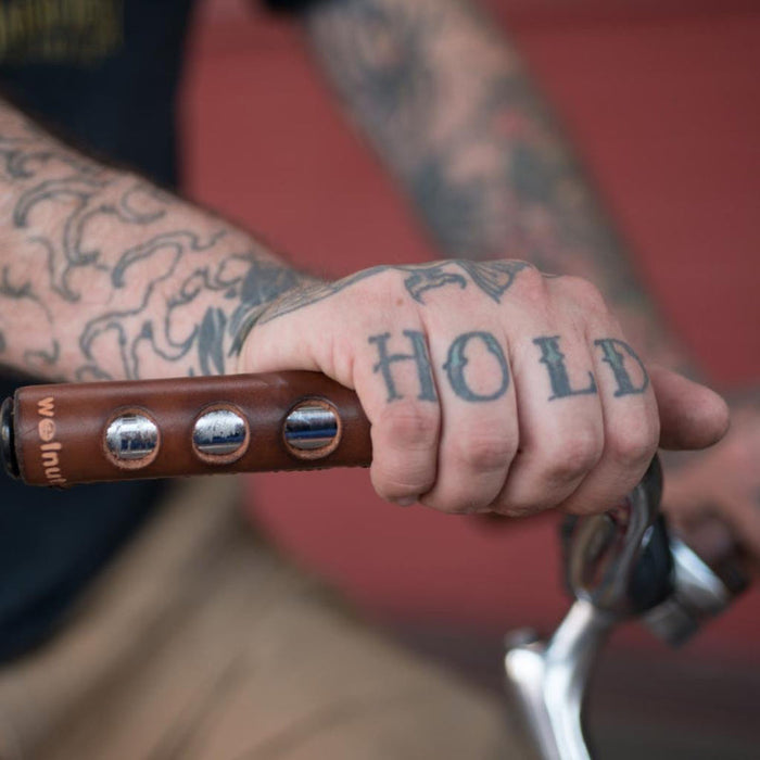 Tattooed man resting his hand on a bicycle city bar wrapped in a dark brown leather grip that has "porthole" round cutouts for finger holds. The man's hand tattoos say "HOLD FAST"