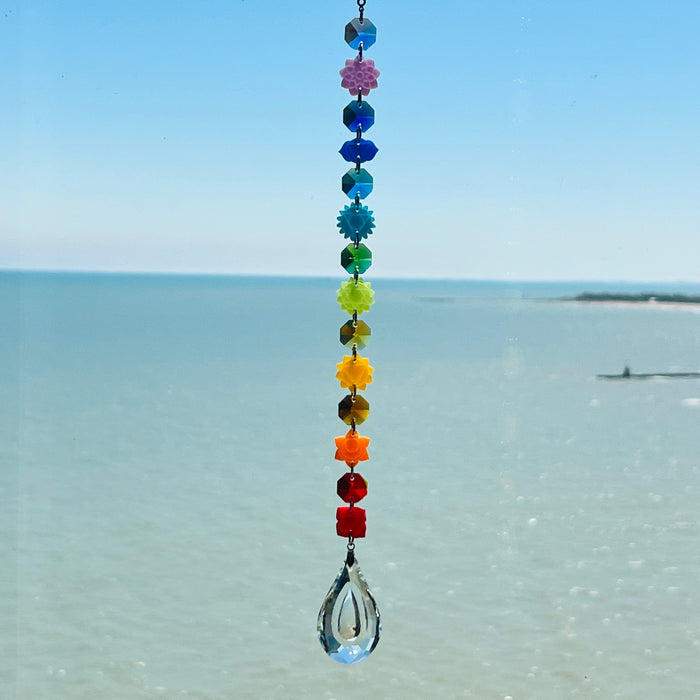 A handmade chakra suncatcher featuring seven stacked polymer clay beads in the colors of the chakras, strung vertically and finished with a hanging faceted glass crystal that reflects rainbows when hit by sunlight. Designed for spiritual, boho, or meditative decor spaces.
