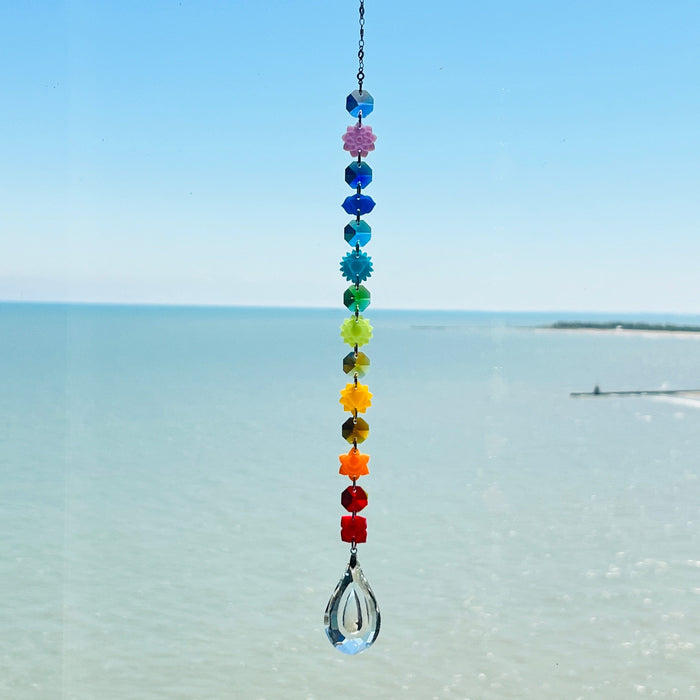 A handmade chakra suncatcher featuring seven stacked polymer clay beads in the colors of the chakras, strung vertically and finished with a hanging faceted glass crystal that reflects rainbows when hit by sunlight. Designed for spiritual, boho, or meditative decor spaces.