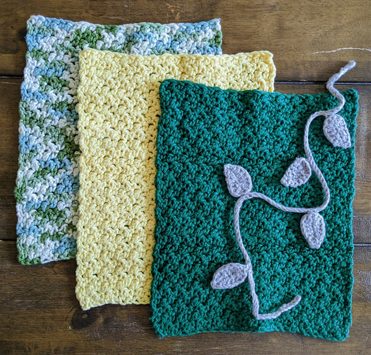 Photograph of a set of three green, yellow, and light blue crocheted kitchen towels overlapping each other, with their grey, leafy crochet tie laid across them.