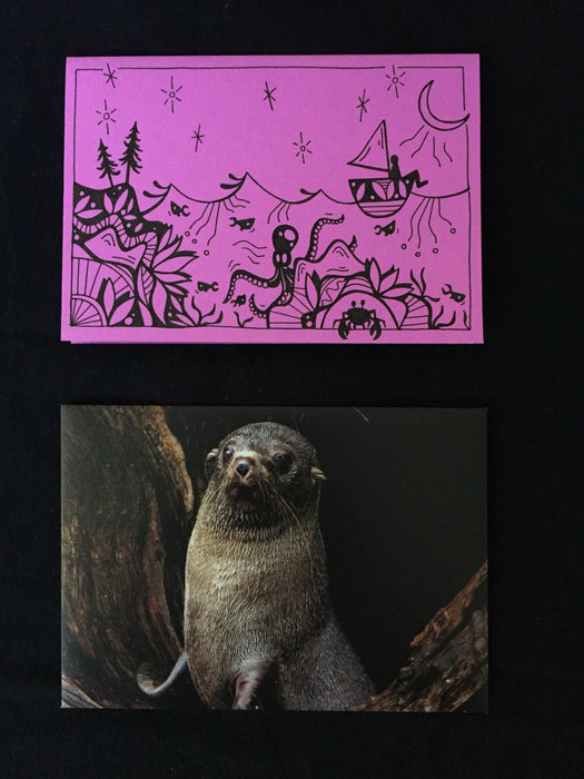 Card features an octopus, crab, and fish swimming below a person on a sailboat under a star-filled night sky on lilac-colored paper. Envelope features photograph of a juvenile sea lion.
