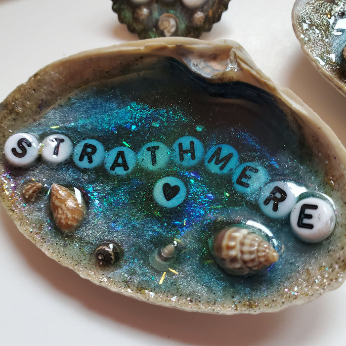 A clam shell featuring a blue resin Beach scene with sand and tiny shells around the outer edge of the shell.  It says Strathmere in the center in white and black letters.