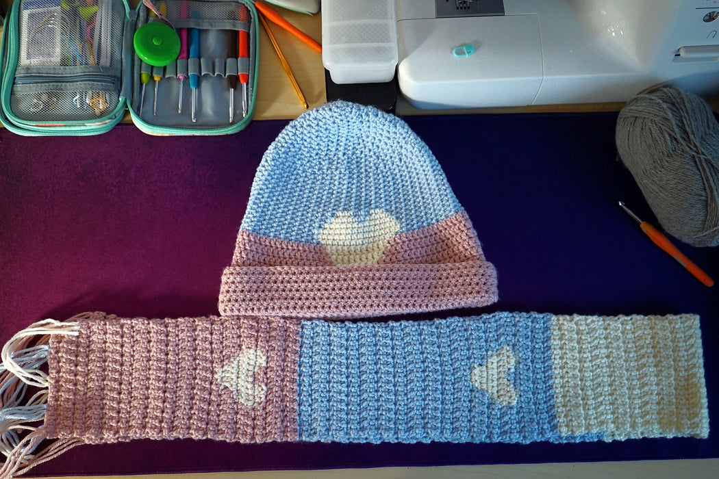 A hat and scarf laid out on a table. The hat's top half is baby blue, the bottom half light pink and folded up. There is a white heart worked into the center, the tip of the bottom just barely covered by the pink. The scarf is made with large sections of pink, blue, and white; it's folded in half. There are two white hearts worked into it, one on the blue and one on the pink. There are loose yarn tassles at the ends.