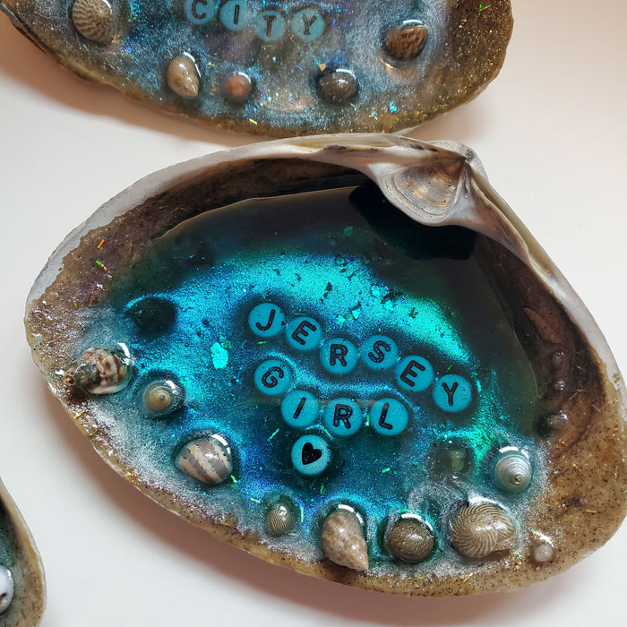 A complete resin Ocean Beach scene is set inside a large natural Clam Shell with the words Jersey Girl in the center using white and black letter beads.  There are tiny shells and Sand at the outer edge to act as the beach area while the blue water is in the center made with resin.