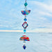 A handmade polymer clay suncatcher featuring marbled rainbow clay pieces in half-circle and quatrefoil shapes, separated by faceted clear glass beads and colorful accents. The suncatcher hangs vertically from a gold chain and includes a central drop-shaped crystal with blue & aqua 14mm octagon crystals. It is photographed in front of a bright blue sky and calm lake horizon, with sunlight highlighting the bright swirls of red, blue, orange, yellow, and green in the clay.