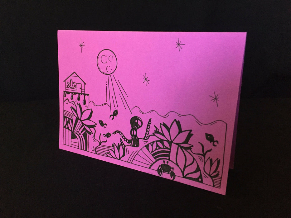 Card features the full moon shining down on an octopus, crab, and fish swimming below a small house on lilac-colored paper.