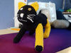 A crocheted cat standing upright. It's mostly black, but its ears and the two legs on our right are yellow, except for the tip of the front paw (which is still black). This one has one mustard yellow button eye and one white button eye. The nose and mouth are white, as are the whiskers, which only this one has.