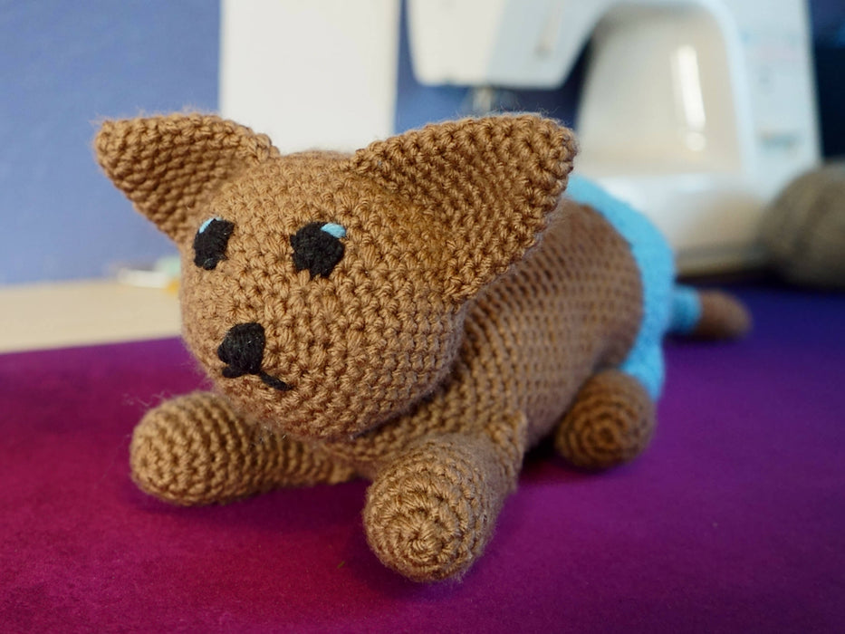 A crocheted cat plushie made to look as if it has brown fur and is wearing blue pants on its butt and hind legs. It's in a crouching pose. Its eyes are embroidered black with light blue highlights.