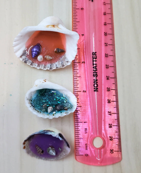 2 ocean resin magnets in natural seashells hung on a metal flower shaped metal stand.  The shells are each colored in a different color, 1 each of bright pink, bright blue, and bright purple.