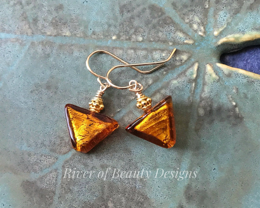 Down-pointing glass triangle earrings in orange and gold, in a green ceramic dish. With ornate little gold accent beads and gold hook ear wires.