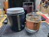 3 glass jars of varying sizes wrapped with leather that is stitched closed with baseball stitch. the smallest jar has neutral-toned leather and black thread; medium jar, dark teal leather and tan-yellow thread; and tallest jar, purple leather with pink thread.