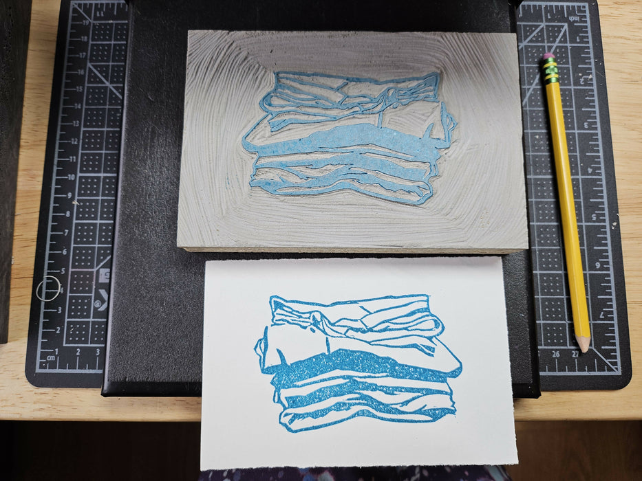 a carved block next to its print; it depict several stacked folded shirts in blue ink