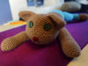 A crocheted cat plushy with green button eyes. The fur is brown, the nose is red, and it's splooting like the white and black cats. This one has larger ears, and it appears to be wearing blue pants on its back half.