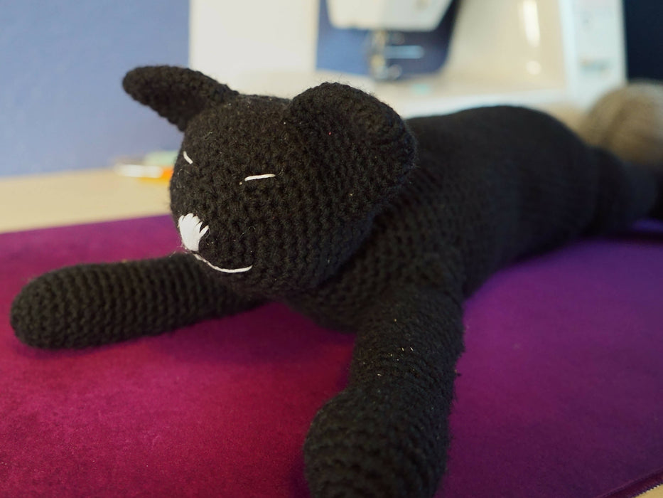 A black crocheted cat with white slits for eyes and a white nose and mouth. Its body is spread out like the small white cat, but its head is held up off the table a bit.