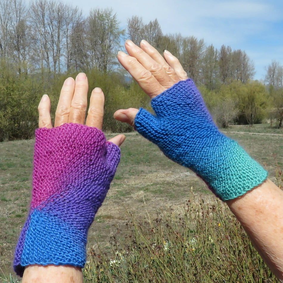 A pair of hands wearing a pair of handmade, fingerless knit gloves. The left glove is a gradient from purple to blue, while the right glove is a gradient from blue to turquoise.