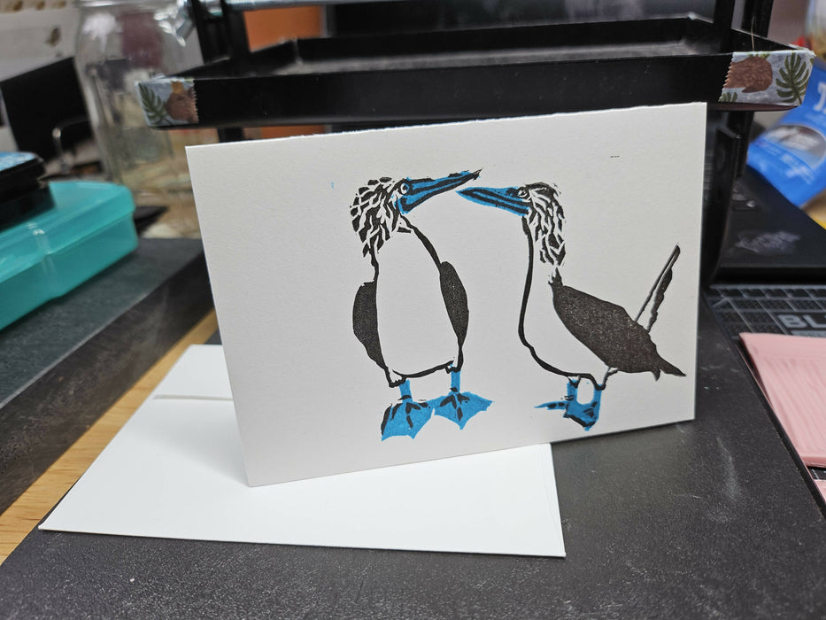 a card propped up on a tiny printing press, next to a plain white envelope. the card has a block print of two blue-footed boobys on it, looking at each other. it's done in two layers of color, black for most of the details and blue for the feet and beaks.