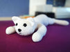 A small crocheted cat plushie shown from the front. Its head and front paws are flat on the table. Its fur is white. Its eyes are embroidered black. One of its back legs can be seen out of focus, spread out behind it.