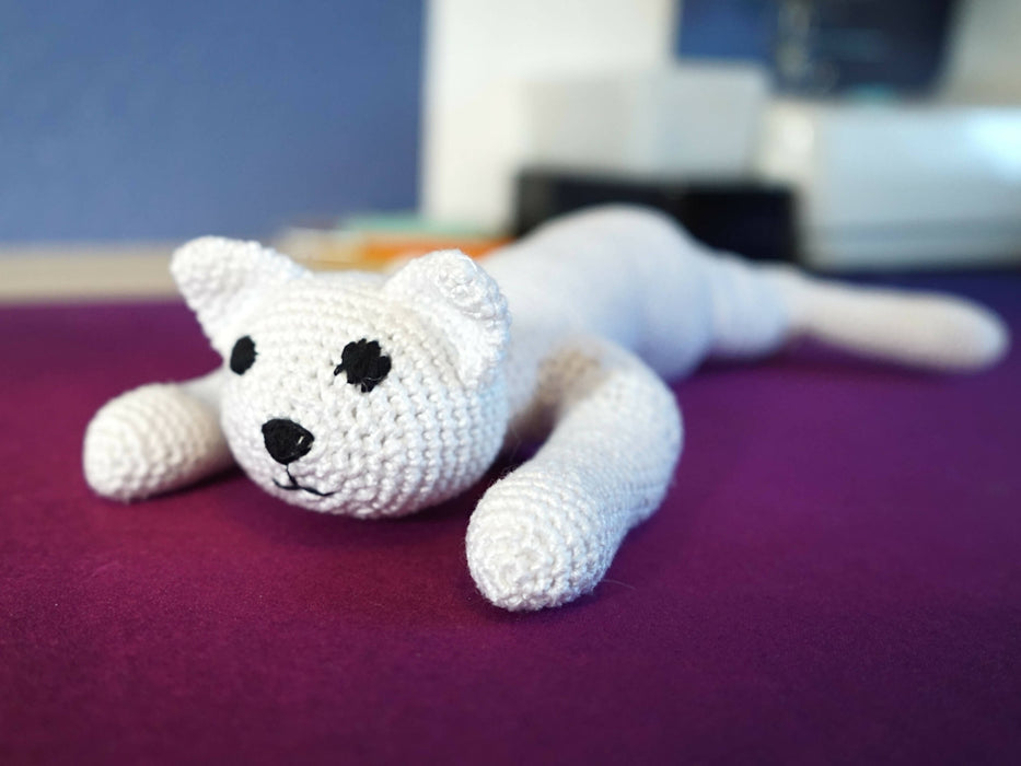 A small crocheted cat plushie shown from the front. Its head and front paws are flat on the table. Its fur is white. Its eyes are embroidered black. One of its back legs can be seen out of focus, spread out behind it.