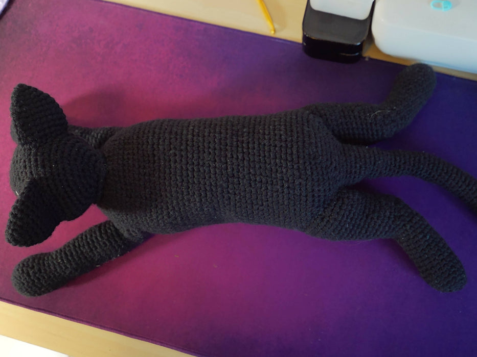 The black cat from the previous image shown from above. Its legs are spread out behind it and its tail is crocheted straight but bendable so it's curved to the side.