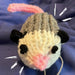 A photo of a tiny knitted opossum. It has black ears, black French knots for eyes, a cute pink tail and a pink nose, and a white face with a gray body