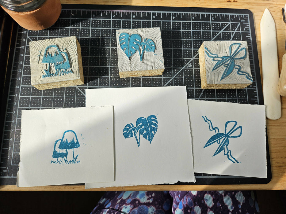 3 carved blocks and their prints: two mushrooms with tall domed caps, some monstera leaves, and sewing scissors snipping a flowing ribbon