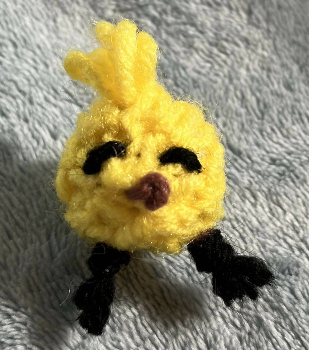 Baldwin the tiny yellow knitted chick. It has a tuft of yellow yarn feathers, a brown yarn beak, and black yarn eyes and feet