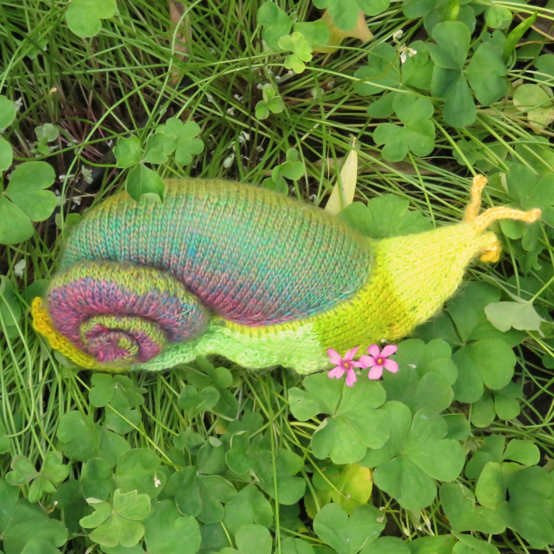 A hand-knit snail stuffed animal in shades of green and purple wool on a bed of moss