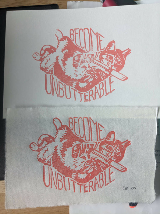 a print in orange ink of a cat laying on its back with paws curled, a butter knife clutched in its mouth. the text reads, in all caps, "become unbutterable"