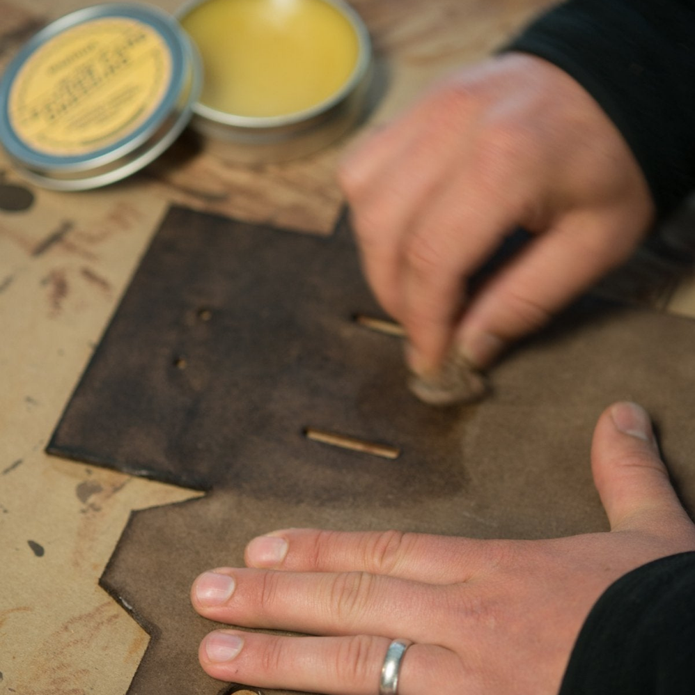 A man rubbing conditioner from a small yellow tin in circular motions on a leather tool on a workbench 