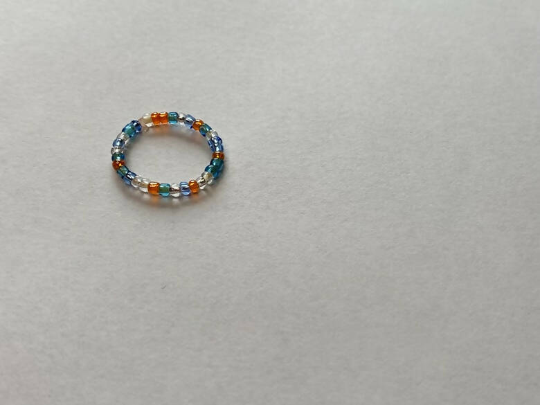 A beaded ring in Artisans Cooperative colors sitting on a gray surface.